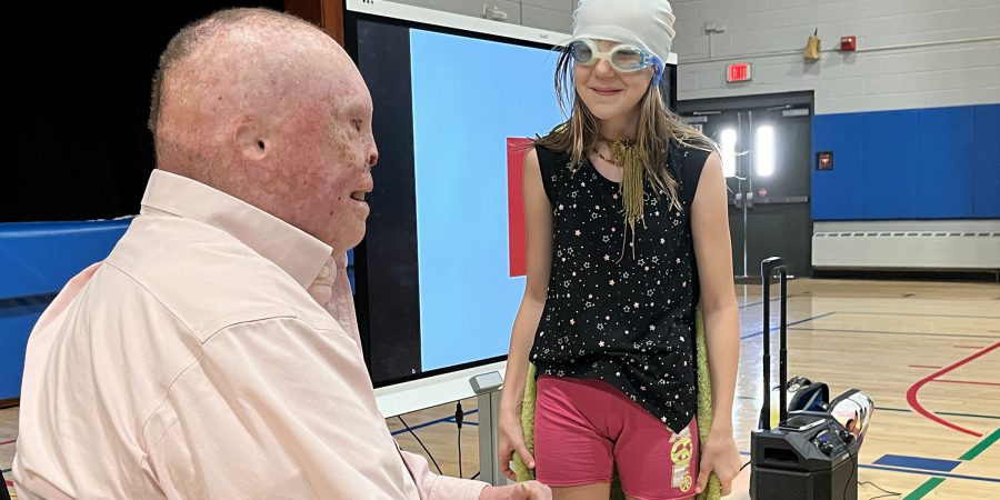 A student with a mask and a cape talks with a disfigured man in a wheelchair