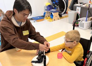 College students helps a kid in a wheelchair write using a device the college kids made