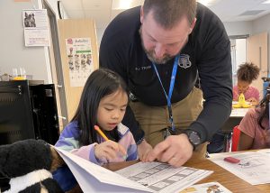 A police officer helps a student in the classroom by pointing to something on her desk