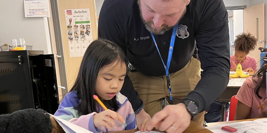 A police officer helps a student in the classroom by pointing to something on her desk