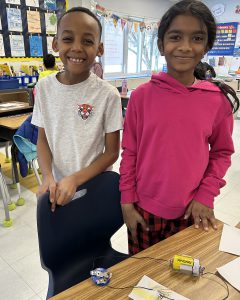 Two students smile while posing in front of an experiment