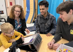 College students and a physical therapist smile while watching a kid in a wheelchair write using a device the college kids made